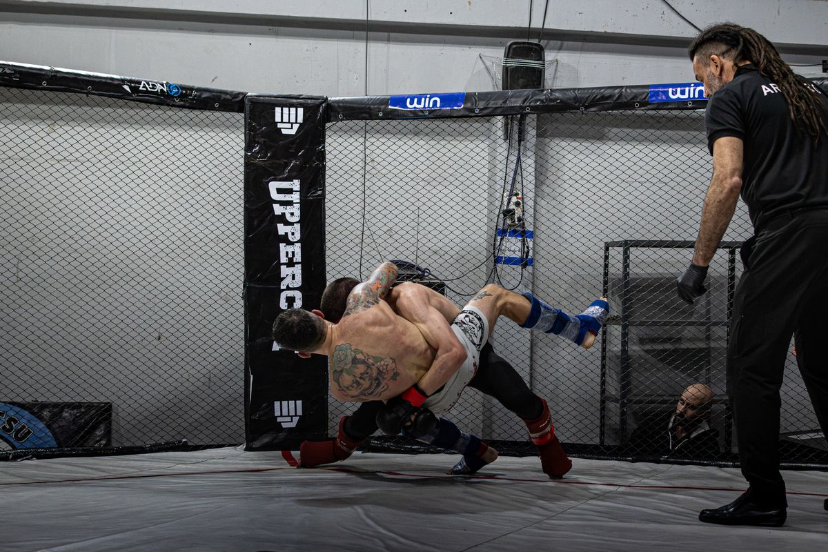Two heavyweight MMA fighters grappling against the cage, one shooting a takedown